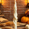 A tall white candle shaped like a mummy or ghost stands on straw and wheat stalks against a brick background.