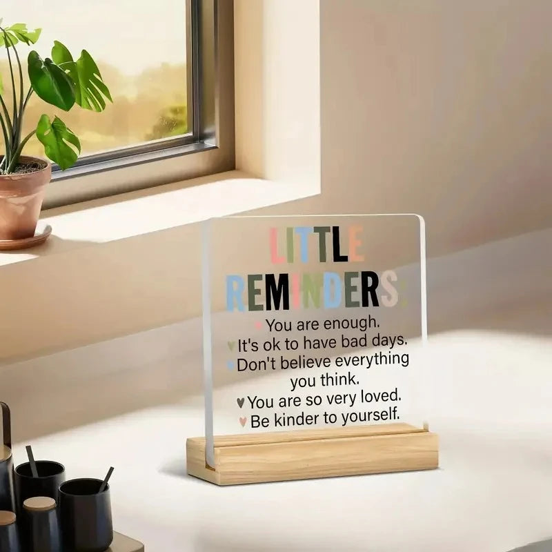 A clear acrylic desk sign with a positive message about self-love and kindness sits on a light surface next to a window.