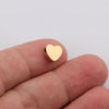 A hand holds a small, gold-colored heart-shaped stainless steel bead charm.