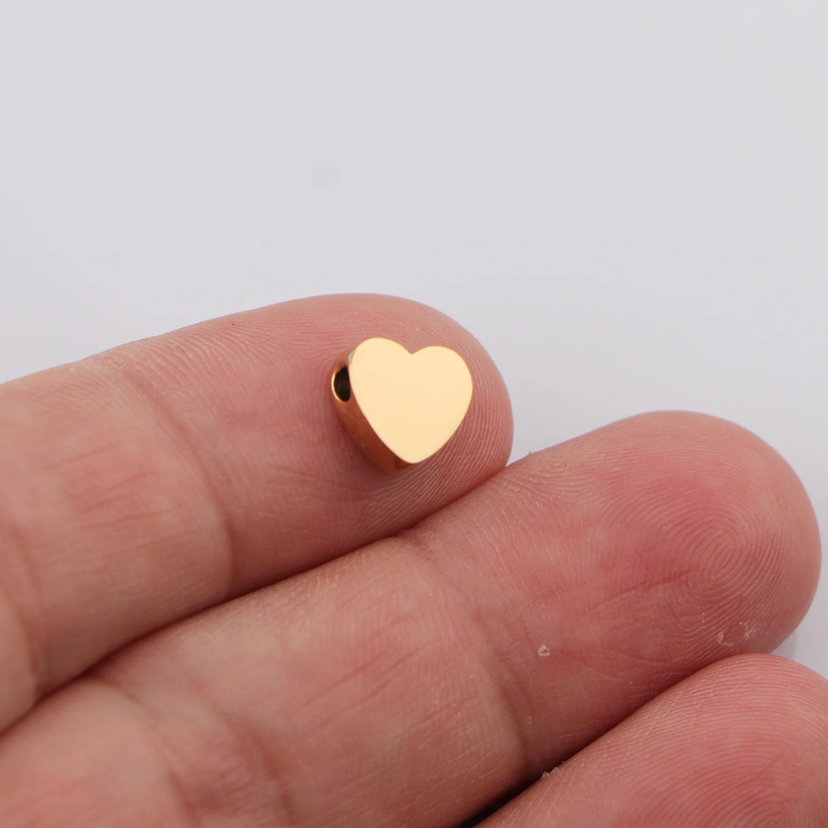 A hand holds a small, gold-colored heart-shaped stainless steel bead charm.