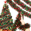 A hand is holding a festive Christmas tinsel garland with red and green branches against a blurred background of a decorated 