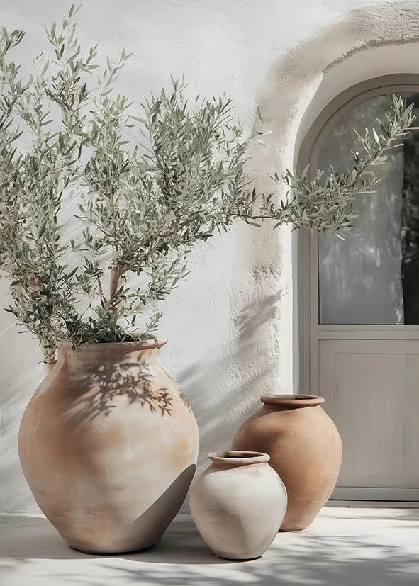A terracotta vase filled with olive branches sits on a white wall next to a light-colored door frame.
