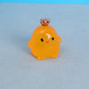 A small, bright yellow resin chicken ornament with a gold crown and black eyes resting on a light blue surface.