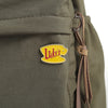 A yellow enamel pin shaped like a smiling face with the word Luke's on it attached to olive green fabric.