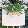 A white sign with gold script welcoming guests, decorated with green eucalyptus leaves and pink roses.