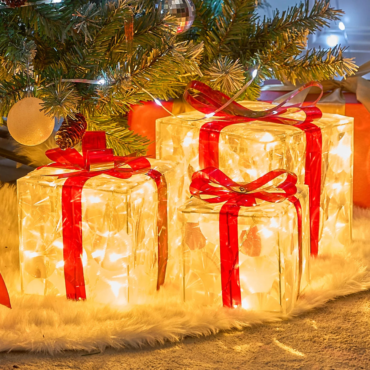 Three clear LED gift boxes wrapped in red ribbons glow brightly on a fluffy surface near a decorated Christmas tree.