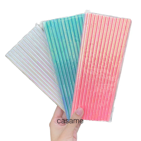 A hand holds three colorful striped paper drinking straws: teal, pink, and iridescent white.