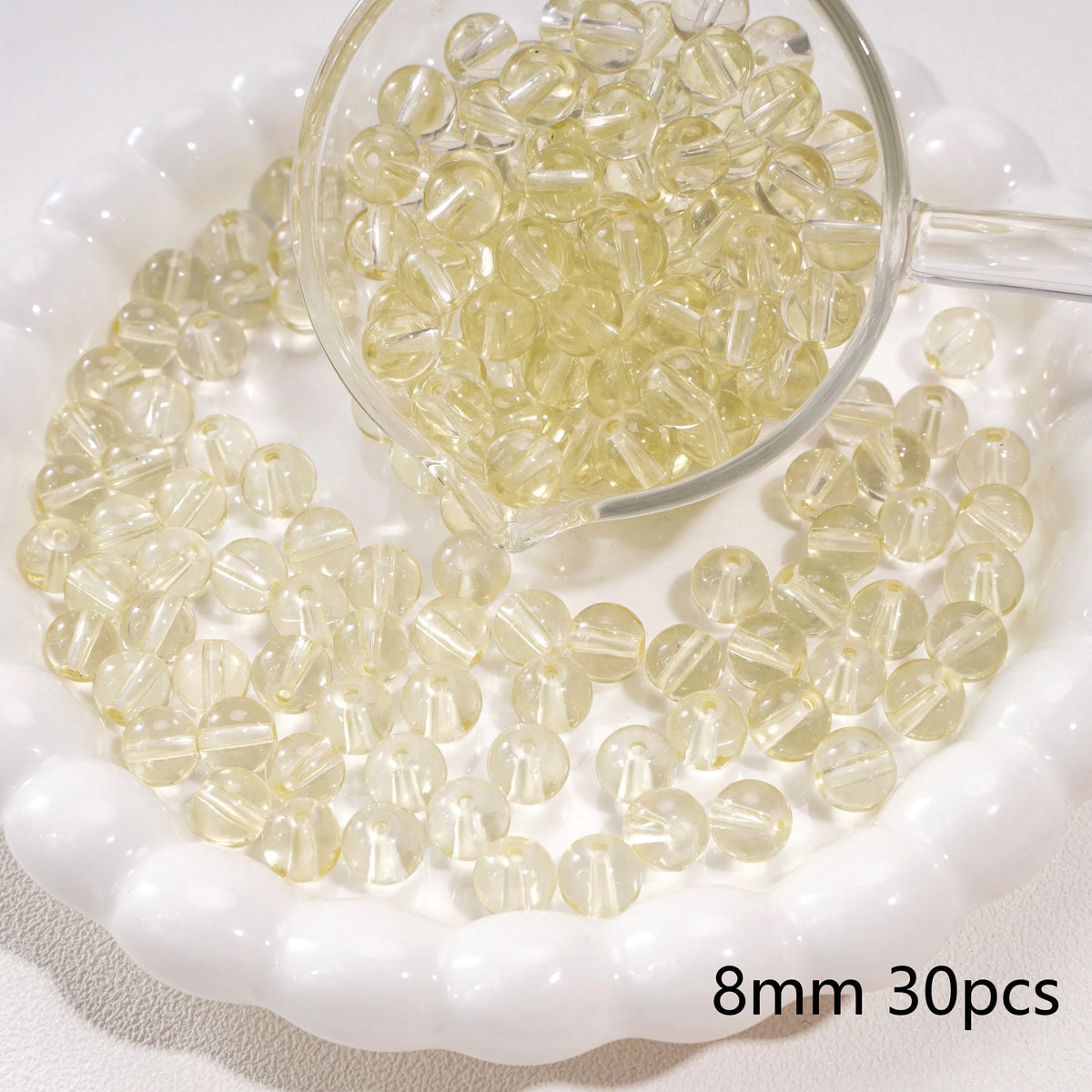 A clear glass measuring spoon holds small round yellow beads scattered on a white dish.