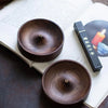 A close-up of a dark brown solid wood incense stick burner resting on an open book with a black box.