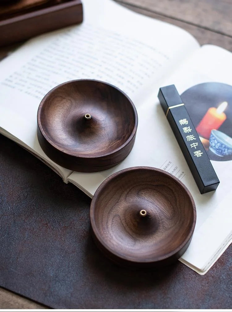 A close-up of a dark brown solid wood incense stick burner resting on an open book with a black box.