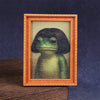 A framed painting of a green frog in a wooden frame.