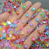 Una mano muestra 50 adornos de uñas en forma de tortuga mini y coloridos esparcidos sobre una superficie.