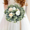 A lush green and cream floral wreath made of roses and eucalyptus leaves, held by a person in a white dress.