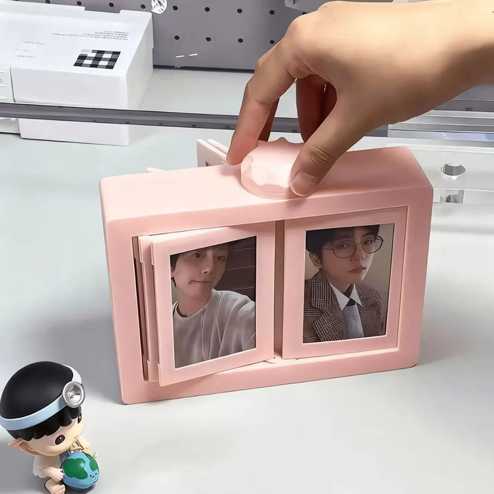 A hand is holding a pink rotating flip photocard display frame with two photo slots, next to a small figurine.