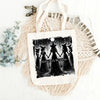 A black and white illustration of four witches standing in a forest with a full moon above them on a white fabric tote bag.
