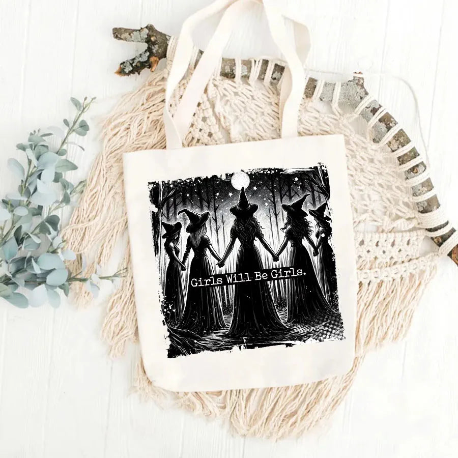 A black and white illustration of four witches standing in a forest with a full moon above them on a white fabric tote bag.