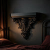 A dark, ornate Gothic wall shelf with intricate carvings mounted on a dark gray wall above a bed.