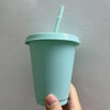 A hand holds a light mint green plastic cold cup with a lid and straw against a plain wall.