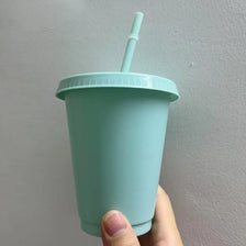 A hand holds a light mint green plastic cold cup with a lid and straw against a plain wall.