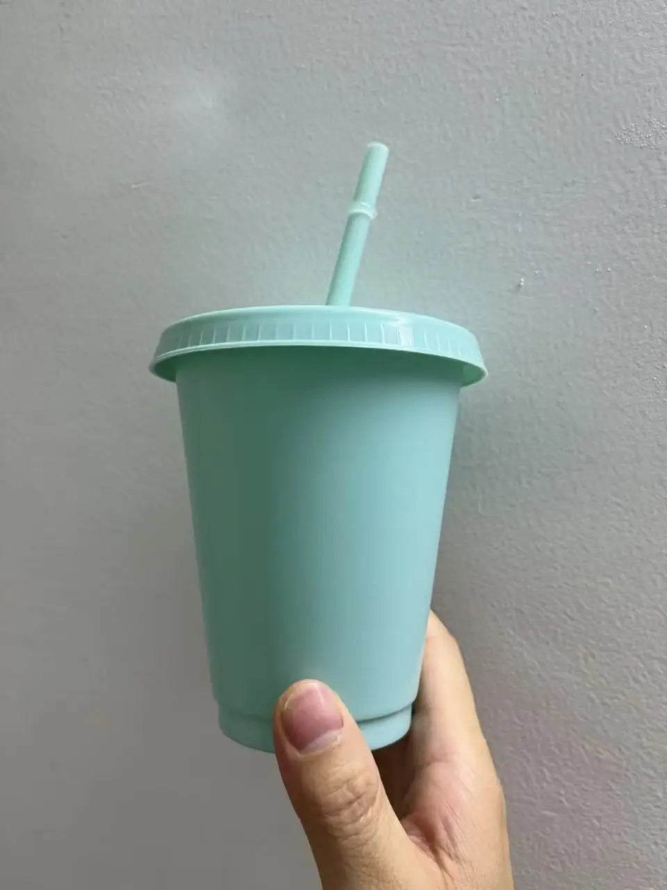 A hand holds a light mint green plastic cold cup with a lid and straw against a plain wall.