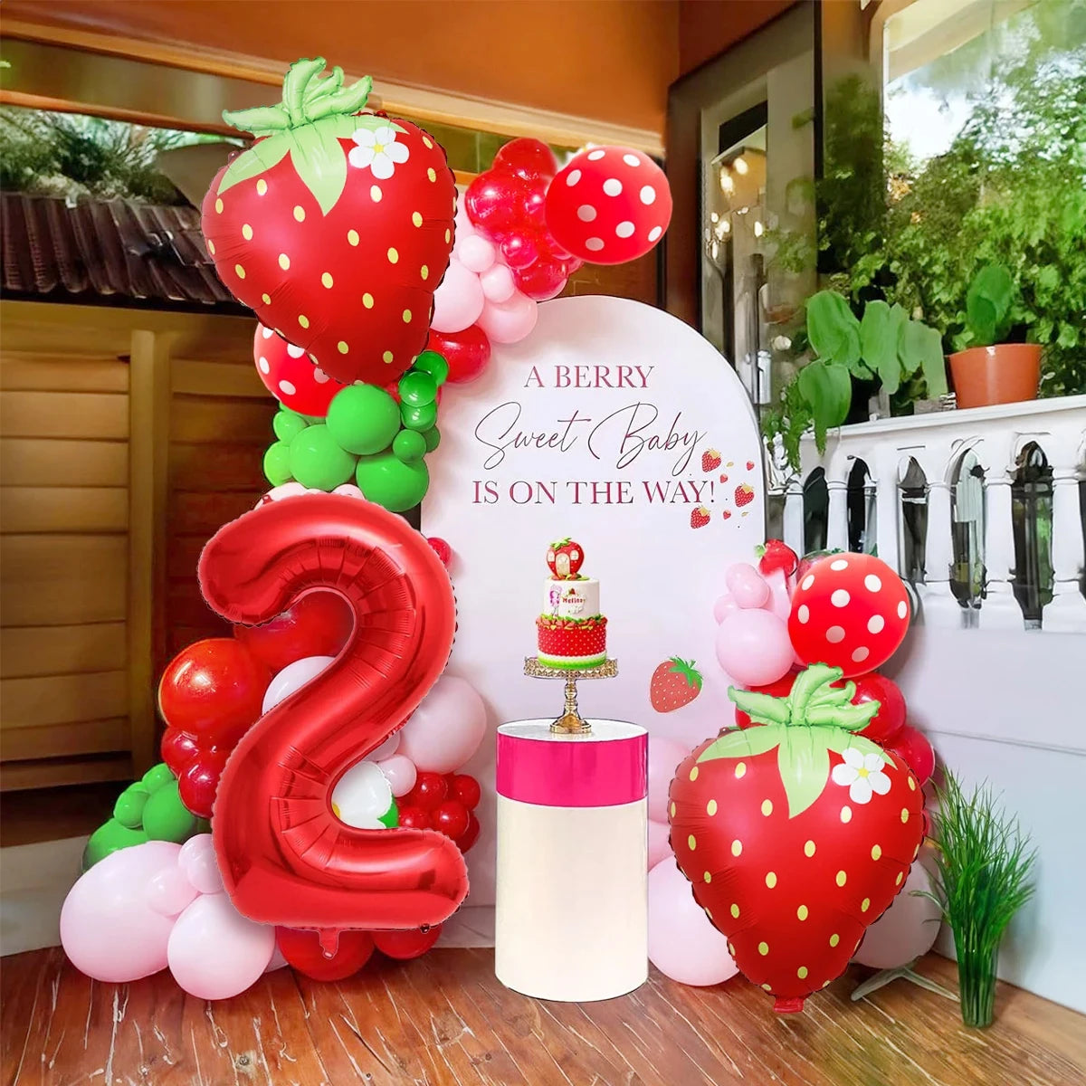 A red and pink strawberry foil balloon set with the number three is displayed on a wooden surface.