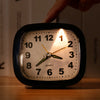 A close-up of a black analog alarm clock with a luminous display showing the time, being touched by a finger.