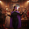 A woman in a black outfit and purple cape with a purple eye mask stands in a richly decorated, dark setting surrounded by oth