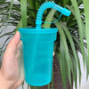 A hand holds a bright blue reusable sipper cup with a silicone straw and lid, set against a background of green foliage.