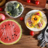 A flat slice of watermelon, a plastic bag of broccoli, and several small red tomatoes arranged on a wooden surface.