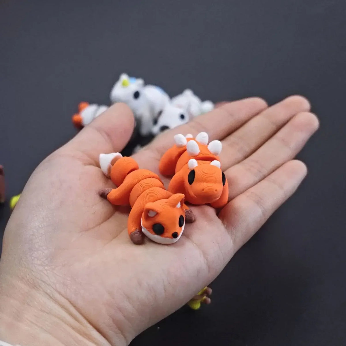 A hand holds several orange and white 3D printed animal figurines, including a fox and a cat.