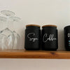 Two black kitchen canisters with wooden lids labeled Sugar and Coffee sitting on a wooden shelf.