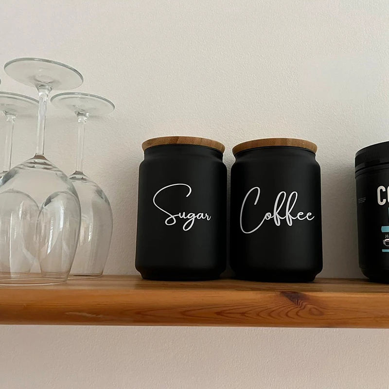 Two black kitchen canisters with wooden lids labeled Sugar and Coffee sitting on a wooden shelf.