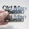 A hand holds two chrome automotive emblems, one reading Old Man Edition and the other reading Old Man Edition.