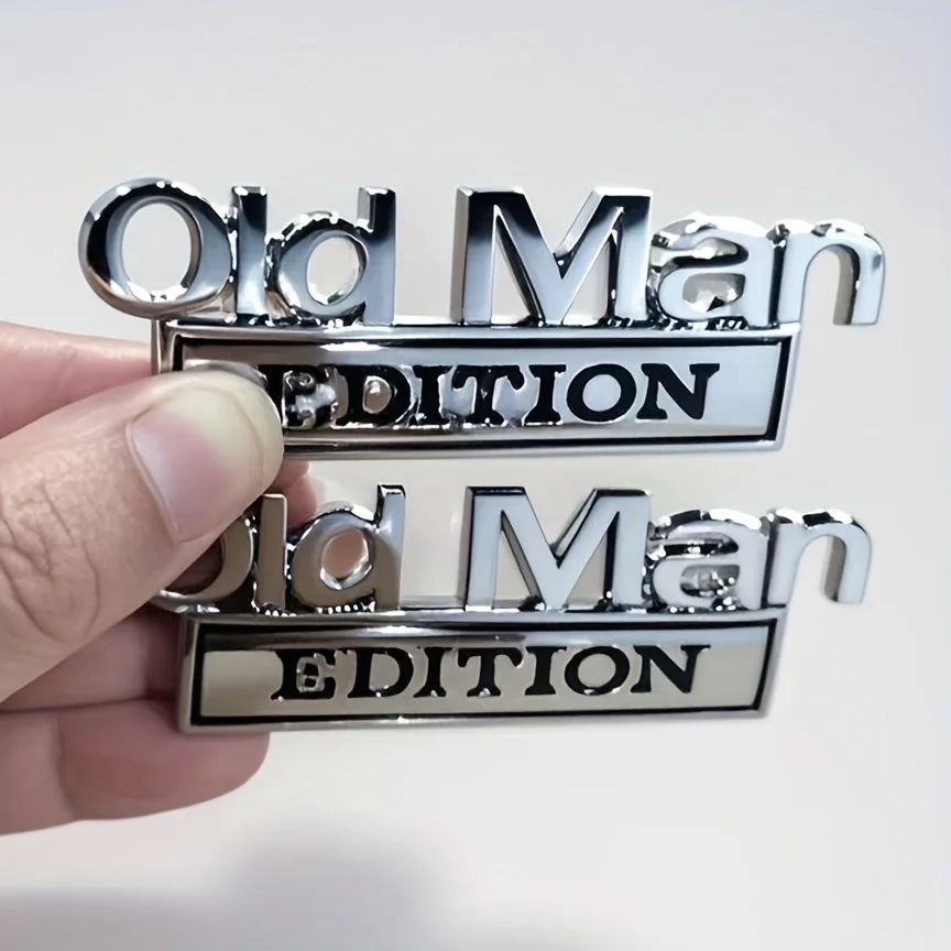 A hand holds two chrome automotive emblems, one reading Old Man Edition and the other reading Old Man Edition.