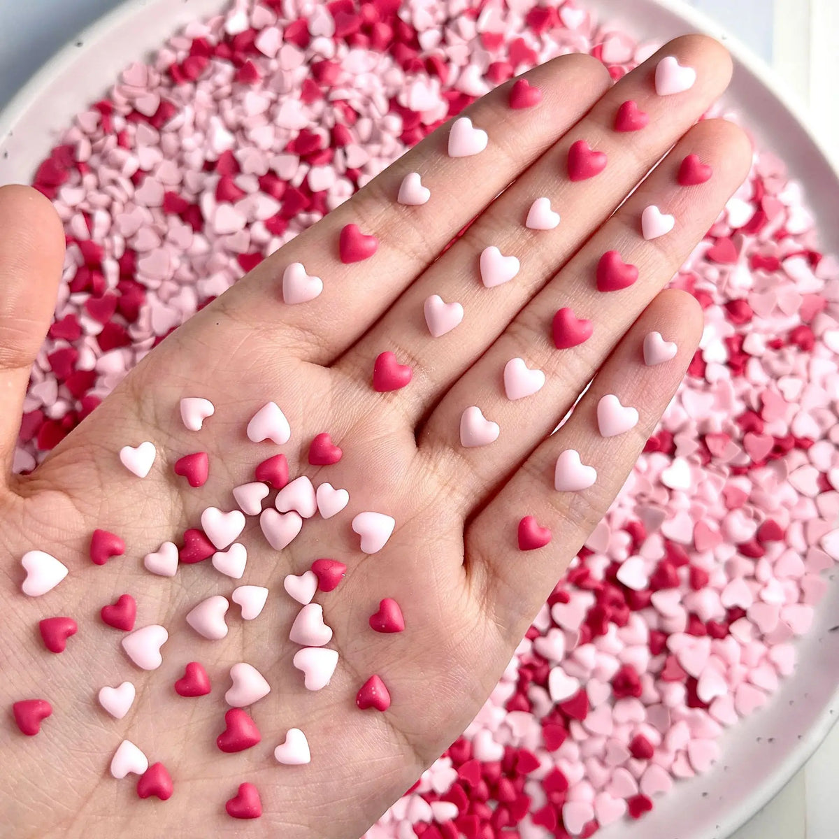 Una mano cubierta de pequeños adornos de uñas con forma de corazón rosa y rojo sobre un fondo rosa.