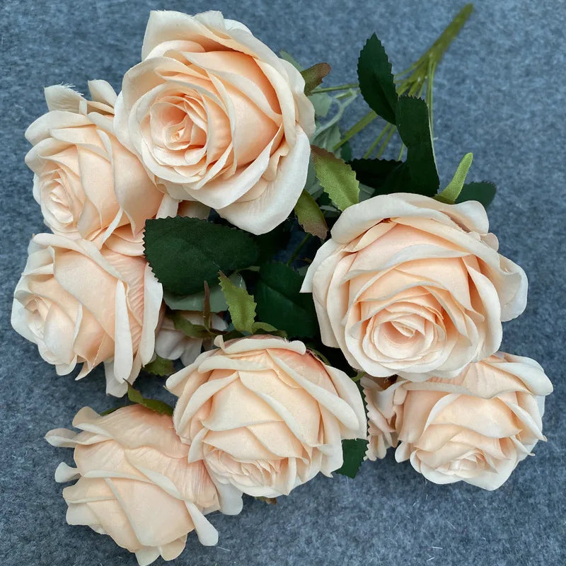 A bouquet of nine realistic silk roses in soft peach and cream tones with dark green foliage.