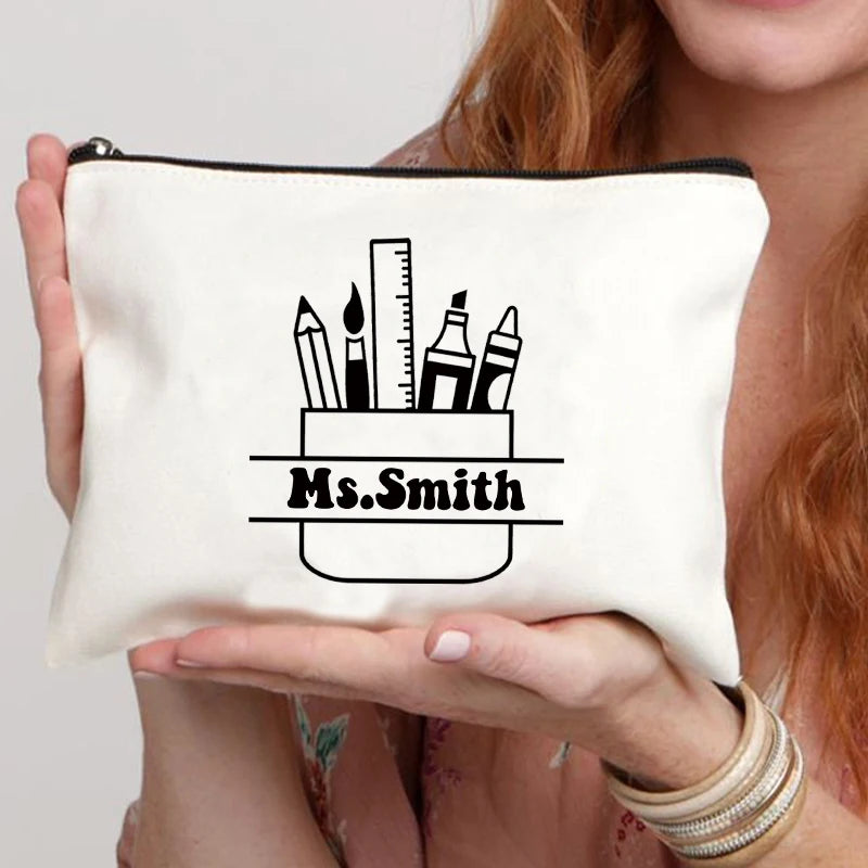 A cream-colored canvas makeup bag featuring a black line drawing of pencils and a name on the front.