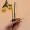 A hand holds several clear glass straws of varying colors and thicknesses next to yellow tulips.
