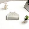 A light gray cat face squeezer tool rests on a white table next to a small glass jar and a potted succulent plant.