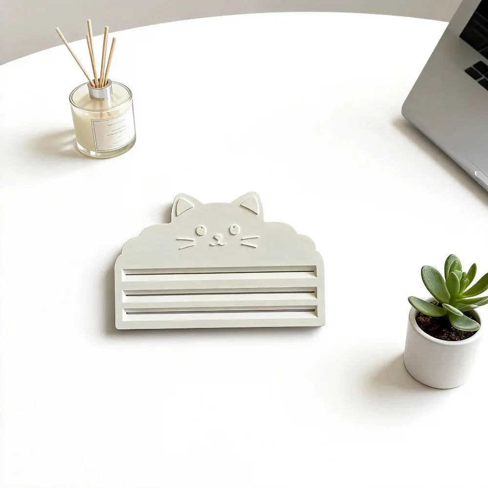 A light gray cat face squeezer tool rests on a white table next to a small glass jar and a potted succulent plant.