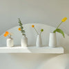 A collection of small yellow and white ceramic flowers in minimalist white vases displayed on a white shelf.