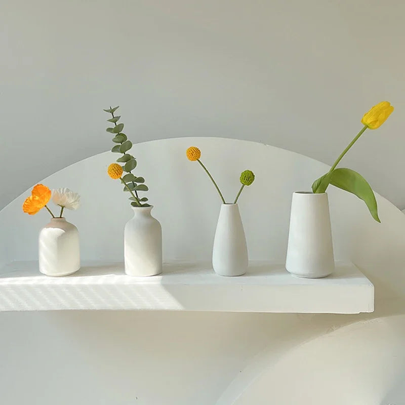 A collection of small yellow and white ceramic flowers in minimalist white vases displayed on a white shelf.