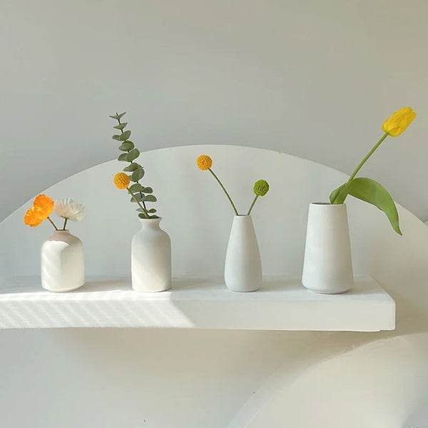 A collection of small yellow and white ceramic flowers in minimalist white vases displayed on a white shelf.