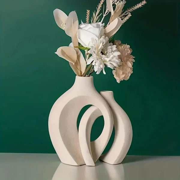 A set of two modern, organic white ceramic vases with hollow designs holding dried floral arrangements against a dark green b