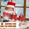 A toddler wearing a Santa hat is holding red festive gift bags in a bright indoor setting.