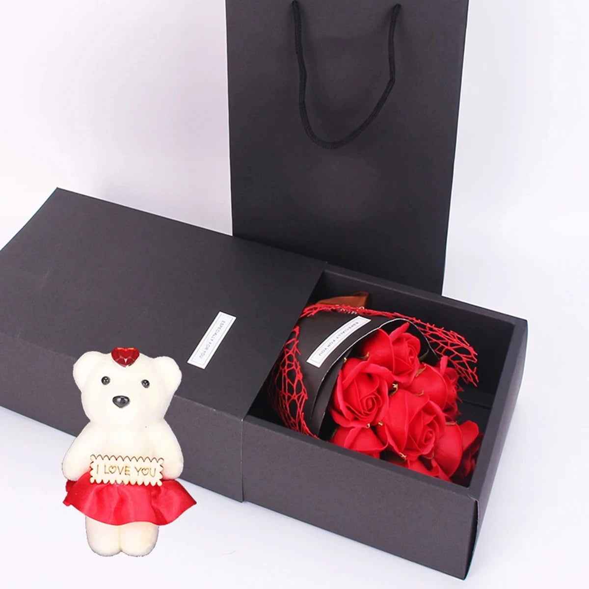Una pequeña figura de oso blanco con un corazón rojo y un ramo de rosas rojas dentro de una caja de regalo negra.