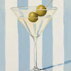 A glass of light yellow liquid with green olives and a wooden skewer, set against a background of vertical blue and white str