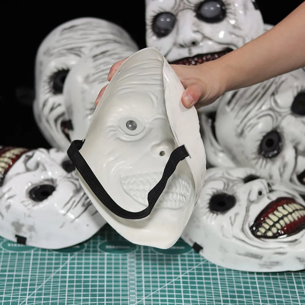 A hand holds a white porcelain demon mask with facial features and teeth, surrounded by other similar masks.