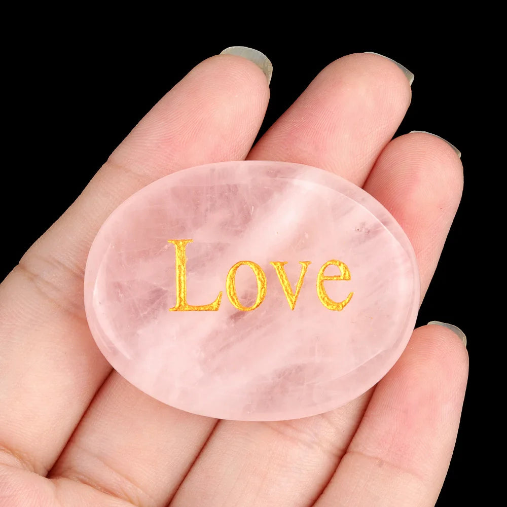 A hand holds a smooth, pink quartz crystal ellipse engraved with the word Love.