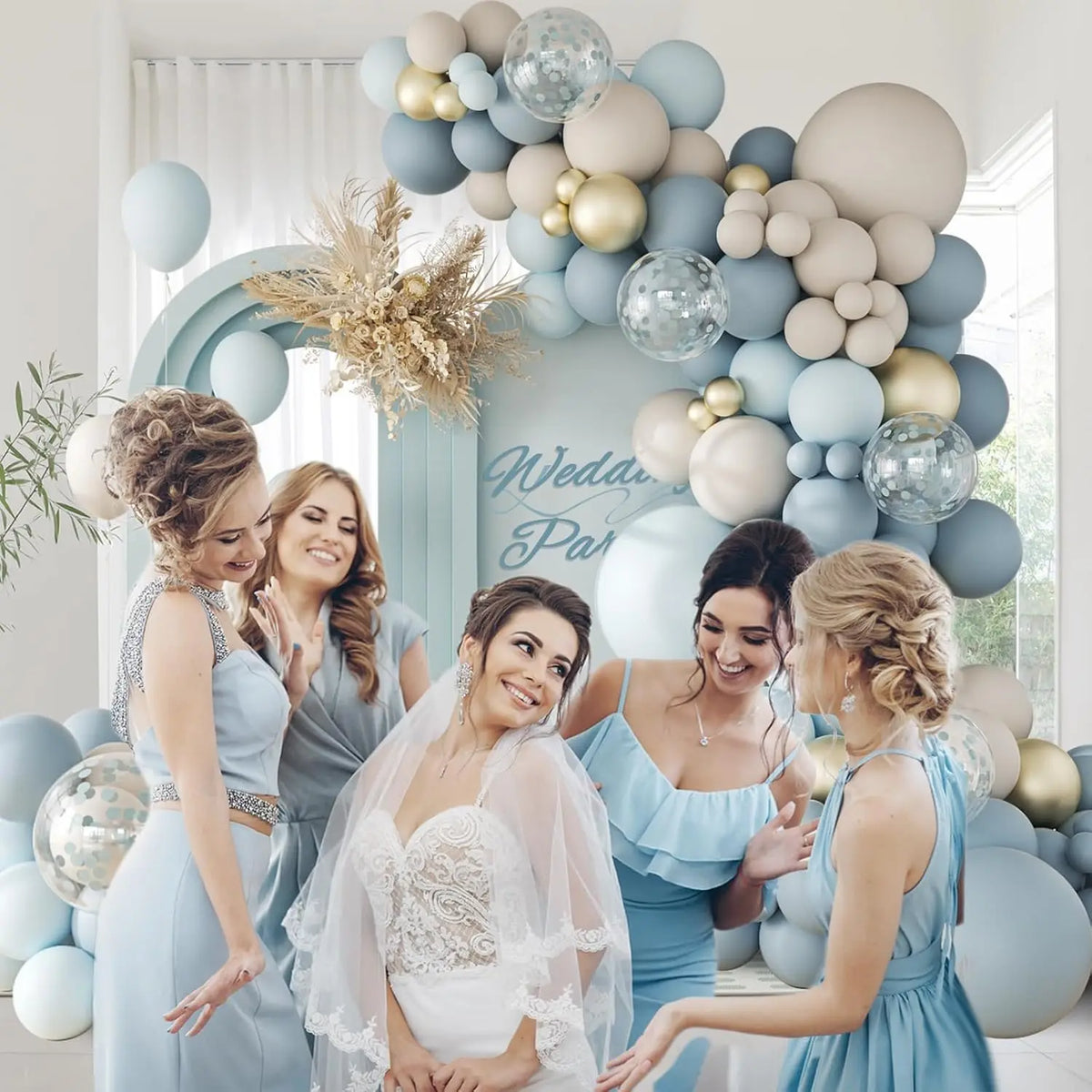 Una novia y damas de honor posan frente a un arco de globos azul claro decorado con globos blancos, grises y dorados.